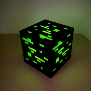 Könnte beinhalten: Eine schwarze würfelförmige Lampe mit einem Muster aus leuchtend neongrünen rechteckigen Ausschnitten auf der Ober- und Seitenfläche. Das Licht strahlt durch die geometrischen Öffnungen und erzeugt einen kontrastreichen visuellen Effekt.
