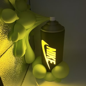 Peut inclure: Une lampe cylindrique en forme de bombe de peinture avec une finition noire mate et un logo Nike jaune lumineux découpé. La lampe est entourée de formes sculpturales vert clair évoquant des bulles, s'étendant de la base et de l'embout.