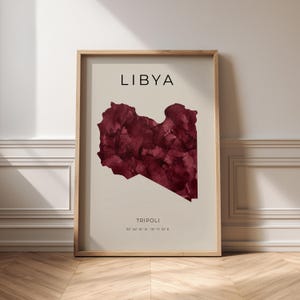 Pode incluir: Uma impressão emoldurada com um mapa da Líbia em aguarela vermelho-escura sobre um fundo bege claro. O texto LIBYA está impresso a preto no topo, com TRIPOLI e as coordenadas 32 52' 31" N - 13 11' 15" E impressas na parte inferior.