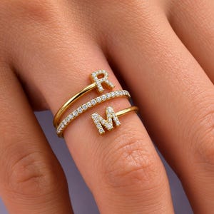 Op de afbeelding: Een goudkleurige wikkelring met de twee hoofdletters R en M, ingelegd met kleine doorzichtige steentjes. De ring bevat een centrale band die ook is bezet met een rij doorzichtige steentjes, wat zorgt voor een gelaagde uitstraling op de vinger.
