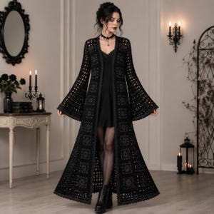 May include: A long, black, open-front crochet duster cardigan featuring a repeating square motif pattern, long flared bell sleeves, and a scalloped hemline, worn over a simple black slip dress with a lace-trimmed neckline and black tights.