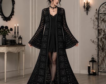May include: A long, black, open-front crochet duster cardigan featuring a repeating square motif pattern, long flared bell sleeves, and a scalloped hemline, worn over a simple black slip dress with a lace-trimmed neckline and black tights.
