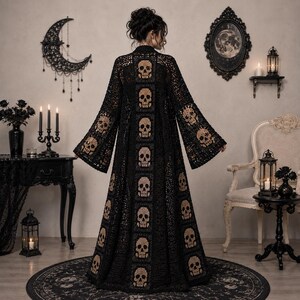 May include: A long, black, open-front crochet duster cardigan featuring a vertical column of beige skull motifs down the center back and additional skull patterns on the wide sleeves. The garment is constructed with an intricate, open-work lace stitch.