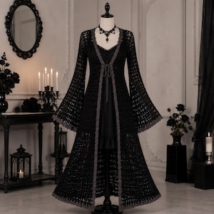 Puede incluir: Un cárdigan largo de ganchillo negro con mangas largas acampanadas, frente abierto con un fino cierre de lazo y un borde de encaje gris festoneado. La prenda se muestra en un maniquí, superpuesta sobre un vestido negro con cuello de encaje.