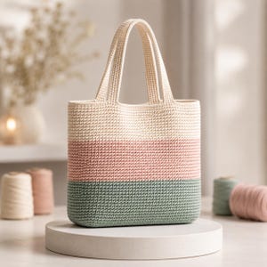 May include: A rectangular crochet tote bag featuring three horizontal color-blocked sections in cream, soft pink, and sage green. The bag has two integrated top handles and a dense, uniform stitch pattern throughout its structured, upright body.