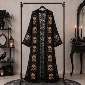 May include: A long, open-front cardigan made of black crochet fabric. The garment features a repeating pattern of beige skull motifs integrated into square panels throughout the body and sleeves, with a delicate lace-like border along the edges.