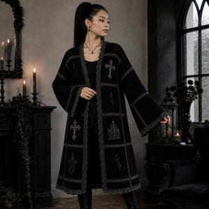 Könnte beinhalten: Eine lange, vorne offene schwarze Strickjacke im Patchwork-Design mit grauen Stickmotiven wie Kreuzen, Spitzbogenfenstern und Sternen. Das Kleidungsstück hat weite Ärmel und einen grauen, wellenförmigen Saum entlang der Kanten und Nähte.