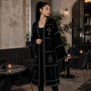 Könnte beinhalten: Eine lange, vorne offene schwarze Strickjacke im Patchwork-Design mit grauen Häkelrändern. Das Kleidungsstück ist mit grauen Stickmotiven verziert, darunter Kreuze, ein gotischer Torbogen, eine Kerze und vierzackige Sterne auf jedem Teil.