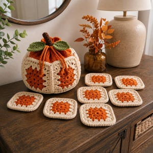 May include: A pumpkin-shaped crochet drawstring pouch featuring orange and cream granny square panels, a brown stem, and two green leaves, accompanied by six matching square crochet coasters in the same orange and cream color scheme.