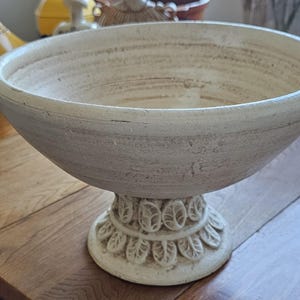 Op de afbeelding: Een crèmekleurige keramische schaal op voet met een brede, open kom en een getextureerde, verweerde afwerking. De voet is voorzien van een decoratieve ring met verhoogde, bladvormige gravures die de stam omringen voor stabiliteit.