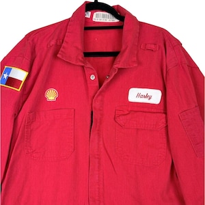 May include: A red long-sleeve work shirt featuring a Texas flag patch on the right sleeve, a Shell logo patch on the right chest, and a white rectangular name patch with the name Harley embroidered in red script on the left chest pocket.