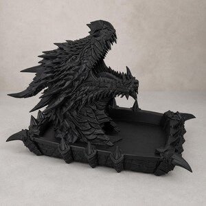 May include: A matte black dragon head sculpture integrated into a rectangular tray. The dragon features detailed scales, sharp horns, and an open mouth with visible teeth. The tray base is bordered by small, pointed spikes along the outer edges.