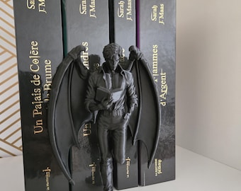 May include: A matte black figurine of a winged humanoid figure holding a book, standing in front of four books by Sarah J. Maas. The figure has large, detailed bat-like wings, a collared shirt, and trousers. The book spines show titles in French.