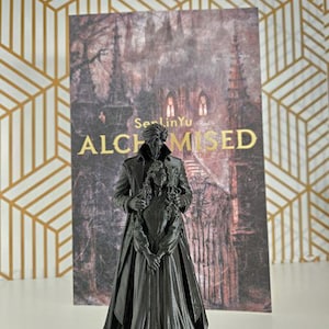 May include: A dark gray 3D printed figurine of a couple standing together, positioned in front of a rectangular print featuring a gothic castle and the text SenLinYu ALCHEMISED in gold lettering. The male figure wears a long coat, and the female figure.