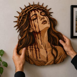 Puede incluir: Una pieza de arte mural de madera en capas que representa una figura con los ojos cerrados y una corona de espinas. La obra presenta varios tonos de veta de madera marrón y gotas brillantes de color rojo intenso que caen por el rostro y cuello.