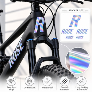 Op de afbeelding: Een matzwart fietsframe met holografische R en ROSE stickers. Een stickerset wordt in de hoek weergegeven met de tekst STICKER SET en ROSE. Een cirkelvormige inzet toont het holografische materiaal. Vijf iconen geven de kenmerken premium vinyl, UV-bestendig, waterdicht, krasbestendig en langdurig en duurzaam aan.