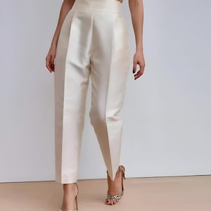 Könnte beinhalten: Hoch taillierte Hose aus cremefarbenem Satin mit glatter, glänzender Oberfläche. Diese maßgeschneiderte Hose verfügt über einen strukturierten Bund, eine klare Vorderseite mit dezenter Faltenlegung und ein schmal zulaufendes Bein.