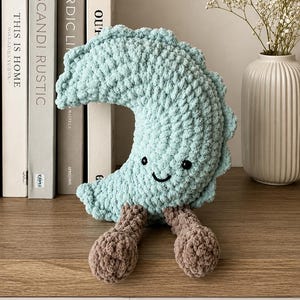 May include: A crescent moon-shaped plush toy made of light blue chunky yarn with a textured surface. It features two small black embroidered eyes, a simple black stitched smile, and two dangling brown legs with rounded feet attached at the base.