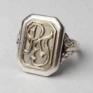May include: A silver signet ring featuring a rectangular face with a textured background and raised, polished gold-toned lettering displaying the initials PS. The ring band has intricate, decorative scrollwork detailing on both sides.