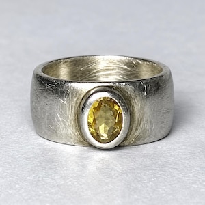 Könnte beinhalten: Ein breiter Silberring mit gebürsteter, texturierter Oberfläche und einem einzelnen oval geschliffenen gelben Edelstein, der in einer glatten, polierten Zargenfassung in der Mitte sitzt. Die Metalloberfläche weist feine Kratzer auf.