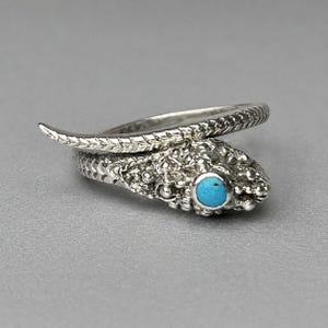 Op de afbeelding: Een zilveren ring in de vorm van een kronkelende slang met een gestructureerd, schubachtig patroon langs de band. De slangenkop is voorzien van een kleine, ronde, felblauwe turkoois als oog, met gedetailleerde metalen afwerkingen.