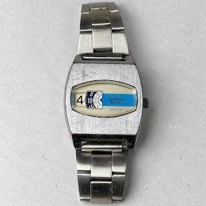 May include: A silver-toned metal digital wristwatch with a brushed finish. The watch face features a horizontal window displaying the time with a blue accent stripe, the text Pallas Eppo, and the word DIGITAL printed in black on a cream background.