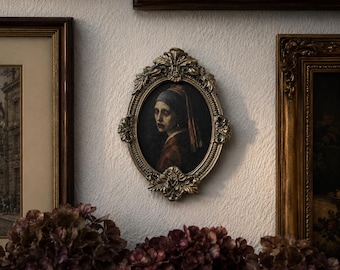 May include: An oval-shaped portrait painting featuring a person wearing a blue headscarf and a red garment, encased in an ornate, antique-style gold frame with intricate leaf and scroll detailing, mounted on a textured white wall surface.