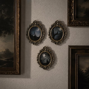 Pode incluir: Três molduras de parede ovais com bordas douradas ornamentadas, cada uma contendo uma ilustração escura e atmosférica de um gato em uma floresta ao luar. As molduras estão dispostas em grupo, rodeadas por arte paisagística retangular.