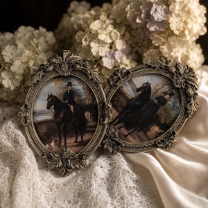 Pode incluir: Dois porta-retratos ovais com detalhes metálicos ornamentados na cor ouro antigo. A moldura esquerda contém uma pintura de uma pessoa a cavalo com um cão, e a moldura direita apresenta uma pintura de uma pessoa montando um cavalo preto.