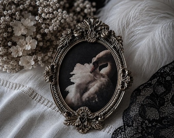 May include: An oval-shaped picture frame with an ornate, antique gold-toned finish featuring intricate leaf and scroll detailing. The frame holds a dark-toned print depicting a white swan with its head tucked into its plumage against a black background.