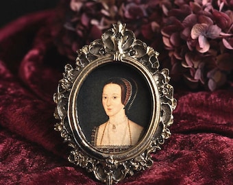 May include: An oval-shaped, ornate metallic picture frame with intricate scrollwork and floral detailing. Inside the frame is a portrait of a person wearing a dark, patterned headpiece and a necklace featuring a prominent letter B pendant.