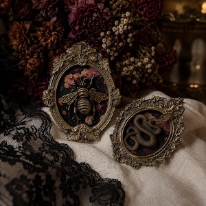 May include: Two oval-shaped decorative frames with ornate gold-toned metal borders. One frame contains a three-dimensional gold bee figure, while the other features a coiled gold snake figure. Both are set against a dark floral fabric background.