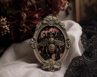 May include: An oval-shaped decorative frame with an ornate, antique gold-toned metal finish. The center features a three-dimensional brass-colored bee mounted against a dark fabric background patterned with small, muted pink floral blossoms.