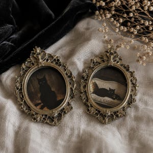 Puede incluir: Dos marcos de fotos ovalados con un acabado dorado metálico ornamentado y detalles de filigrana intrincados. Cada marco contiene una pequeña impresión de un gato negro; uno muestra un gato sentado y el otro un gato tumbado en un sofá.