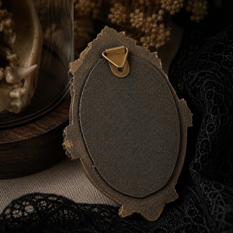 May include: An oval-shaped picture frame with a dark bronze-colored finish and an ornate, textured border. The back of the frame features a solid dark brown backing material and a triangular metal hanging hook attached at the top center.