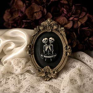 Può includere: Una cornice ovale decorata color oro contenente un cammeo nero. Il cammeo presenta un'illustrazione bianca di due scheletri congiunti con un cartiglio sottostante che riporta il testo EMEER ET UIRUE in lettere maiuscole nere.