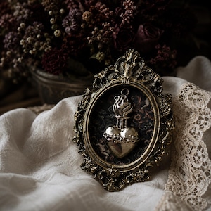 May include: An oval-shaped wall hanging featuring an ornate, antique gold-toned metal frame. Inside the frame is a dark, patterned background with a central silver-toned metal heart emblem, which is topped with a stylized flame and encircled by thorns.