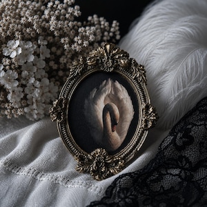 Pode incluir: Uma moldura oval com um acabamento dourado antigo ornamentado, apresentando detalhes intrincados de folhas e volutas. A moldura contém uma impressão escura de um cisne branco com as asas arqueadas para cima, com um bico preto e toques vermelhos.