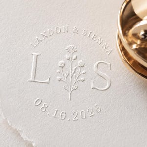 May include: A wax seal impression on textured off-white paper featuring the letters L and S, a central floral sprig, and the text LANDON & SIENNA 08.16.2026. A gold-colored metal wax seal stamp is positioned at the top right of the paper.