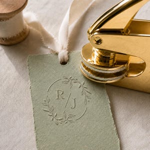 May include: A gold metal embosser tool pressing onto a rectangular sage green paper tag. The tag features a debossed circular laurel wreath design surrounding the letters R and J separated by a forward slash. A wooden spool with white ribbon is visible.