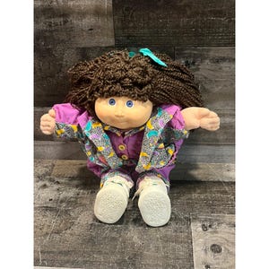 Könnte beinhalten: Eine Puppe mit braunen Wollhaaren, blauen Augen und heller Haut. Die Puppe trägt einen violetten Overall mit einer gemusterten Jacke in Gelb, Türkis und Schwarz. Sie hat weiße Turnschuhe mit Schnürsenkeln und ein türkisfarbenes Haarband.