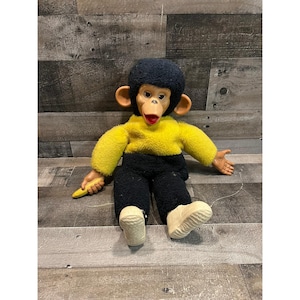 May include: A plush monkey doll with a black fuzzy head, a bright yellow fuzzy torso, and black fuzzy pants. The doll has a tan plastic face with large ears, a red mouth, and tan plastic hands. It holds a small yellow plastic banana in one hand.