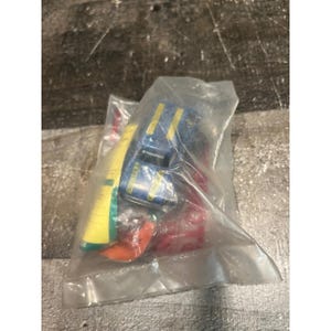 May include: A blue toy truck with yellow stripes and the word STOMPER printed on the hood, enclosed in a clear plastic bag. The toy is accompanied by a yellow and green plastic component and a red plastic piece, all sealed within the packaging.