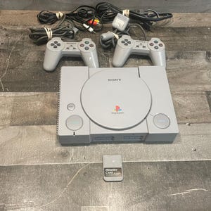 May include: A gray Sony PlayStation game console with two matching gray controllers, a gray memory card, and associated power and AV cables. The console features a circular disc cover with the Sony PlayStation logo and buttons labeled Reset and Open.