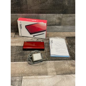 May include: A red Nintendo DS Lite handheld gaming console with its original box, a gray power adapter with a coiled cord, and a white instruction manual. The box features the Nintendo DS Lite logo and an image of the red console.