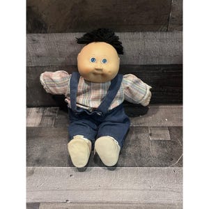 Könnte beinhalten: Eine Puppe mit einem runden, pfirsichfarbenen Gesicht, blauen Augen und kurzem schwarzem Wollhaar. Die Puppe trägt ein langärmeliges kariertes Hemd und eine dunkelblaue Jeanslatzhose mit Trägern sowie einfache, helle Stoffschuhe.