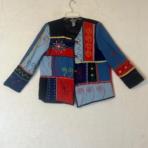 May include: A long-sleeved jacket featuring a patchwork design in red, blue, and black. The garment includes embroidered yellow, red, and blue spiral and star patterns, a V-neckline, and three toggle closures down the front centre on a black trim.