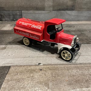 May include: A red and white Coca-Cola tanker truck toy with a cylindrical tank featuring the text First Tank Car Equipped Dispensing and Ice Cold Coca-Cola Served here. The vehicle has cream-colored wheels, an open cab, and a black steering wheel.