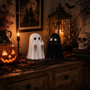 Peut inclure: Deux figurines de fantômes lisses et arrondies, l'une en blanc uni et l'autre en noir uni, chacune dotée de deux yeux sombres et de petits bras levés pour montrer un geste du doigt d'honneur. Ces objets décoratifs sont placés côte à côte.