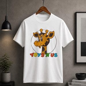 Pode incluir: Uma t-shirt branca de manga curta com um gráfico de uma cabeça de girafa de desenho animado dentro de um círculo preto fino. Abaixo da girafa, o texto TOYS R US está impresso em letras coloridas com uma textura de estilo vintage.